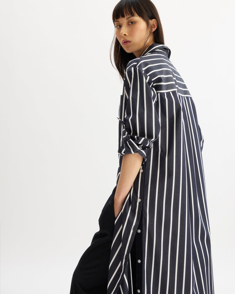 A woman with straight dark hair models the Arlo Mott Lafayette Side Button Maxi Dress in Navy Stripe, crafted from Portuguese poplin, featuring rolled-up sleeves, as she looks over her shoulder against a white background.