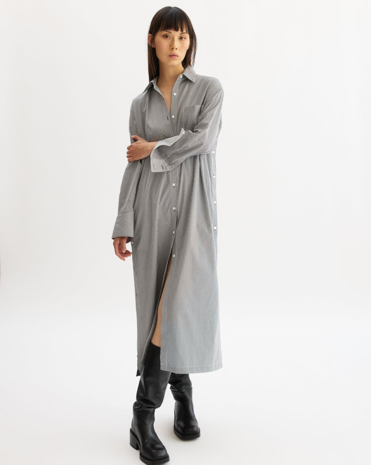 A person stands against a plain white background wearing the Arlo Mott Lafayette Side Button Maxi Dress in White Stripe with side pockets and subtle stripes, paired with black knee-high boots. Their arms are crossed and their expression is neutral.
