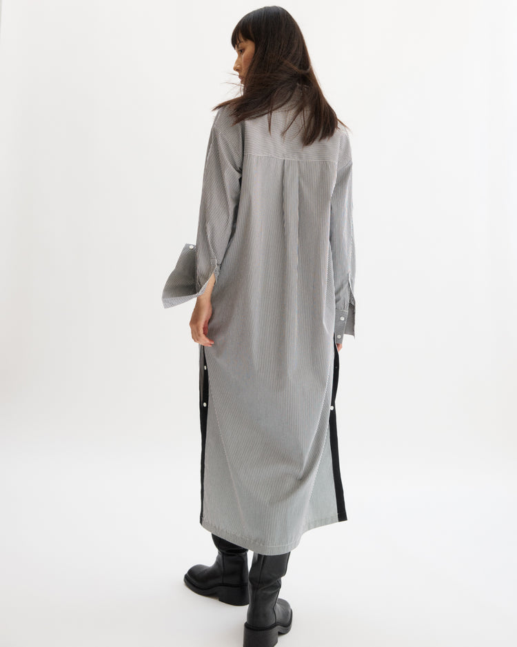 A woman with long dark hair stands facing away, wearing the Arlo Mott Lafayette Side Button Maxi Dress in White Stripe with side pockets and black boots, set against a plain white background.