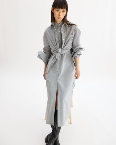 A woman wears the Arlo Mott Lafayette Side Button Maxi Dress in White Stripe, tied at the waist with rolled sleeves and side pockets, paired with black knee-high boots against a plain white background.