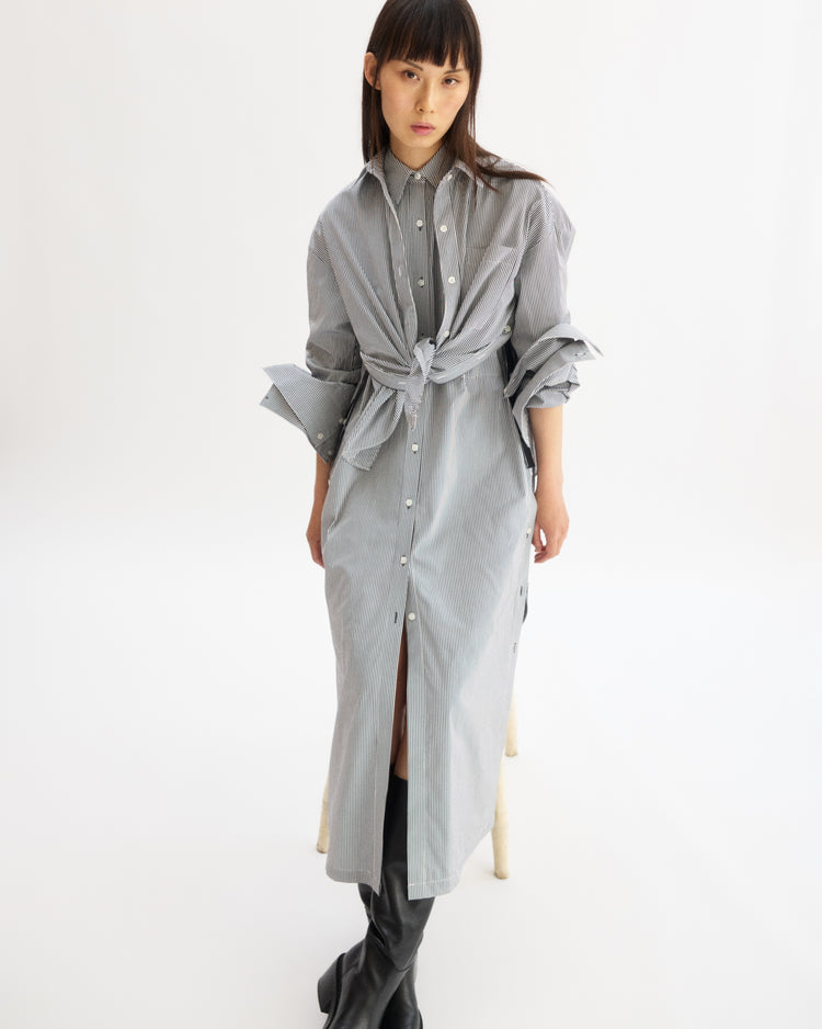 A woman wears the Arlo Mott Lafayette Side Button Maxi Dress in White Stripe, tied at the waist with rolled sleeves and side pockets, paired with black knee-high boots against a plain white background.