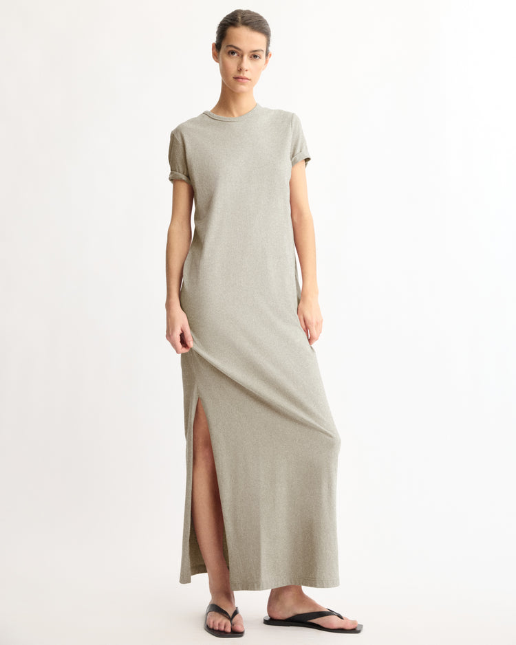 A woman stands against a plain background wearing the Arlo Mott Hubert Short Sleeve Maxi Dress in Grey with black flip-flops. Her hair is tied back, and she looks at the camera with a neutral expression.