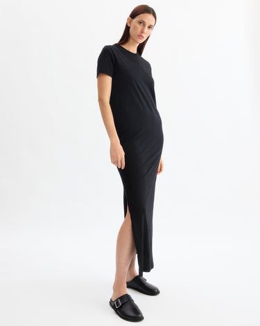 A woman stands on a white background wearing the Arlo Mott Hubert Short Sleeve Maxi Dress in Black, an ankle-length Supima cotton dress with a side slit. She pairs it with black slip-on buckle shoes, has straight brown hair, and a neutral expression.