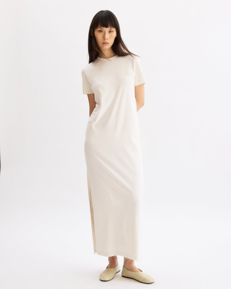A woman with straight, dark hair and bangs stands against a white background, wearing the Arlo Mott Hubert Short Sleeve Maxi Dress in Chalk and beige slip-on shoes. Her arms are behind her back.