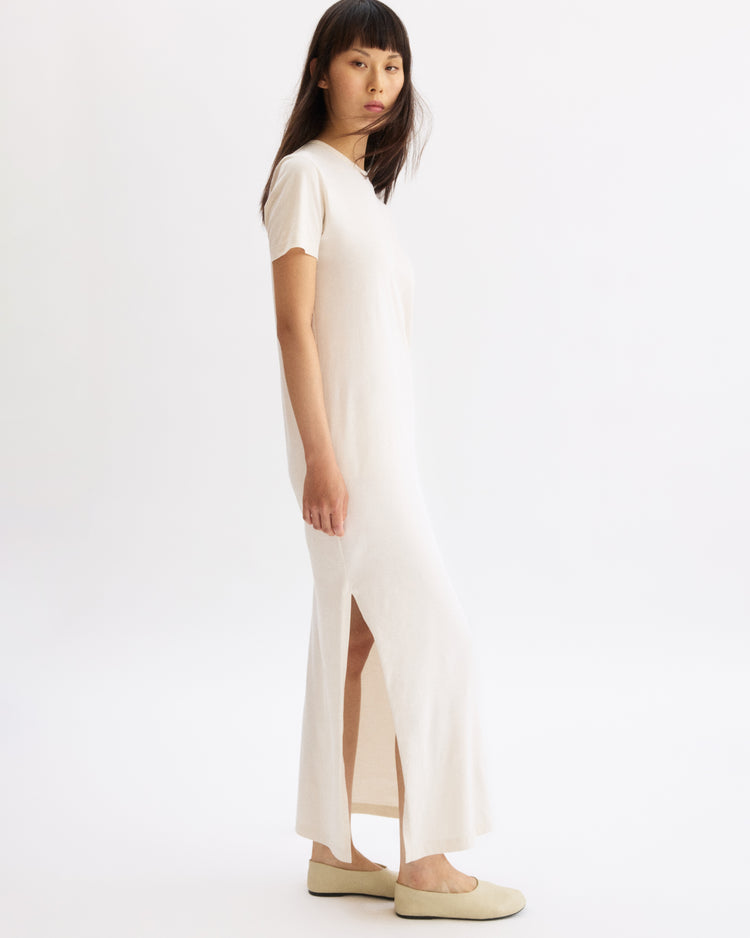 A woman stands sideways against a plain white background, wearing the Arlo Mott Hubert Short Sleeve Maxi Dress in Chalk with light beige flats.