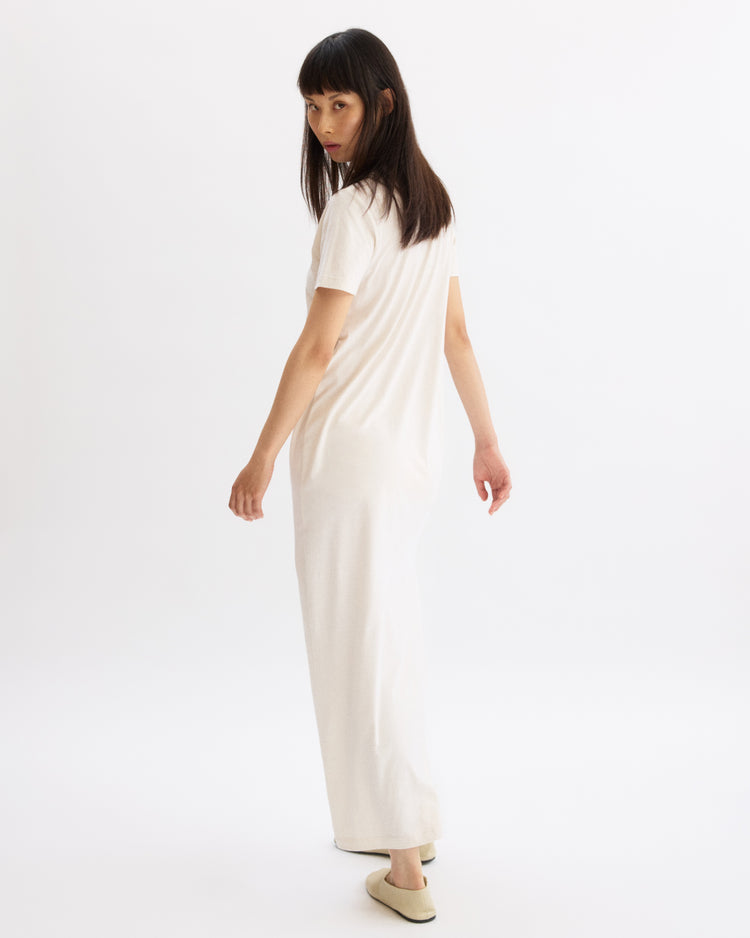 A woman with straight dark hair, wearing the Arlo Mott Hubert Short Sleeve Maxi Dress in Chalk and cream slippers, stands slightly turned away, looking over her shoulder against a plain white background.