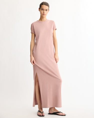 A woman stands against a white background wearing the Arlo Mott Hubert Short Sleeve Maxi Dress in Dusty Rose with a side slit and black flip-flops. Made in New York, she has her hair tied back and looks calmly at the camera.