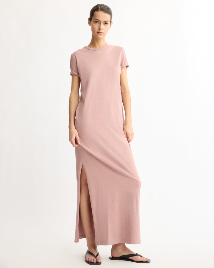 A woman stands against a white background wearing the Arlo Mott Hubert Short Sleeve Maxi Dress in Dusty Rose with a side slit and black flip-flops. Made in New York, she has her hair tied back and looks calmly at the camera.