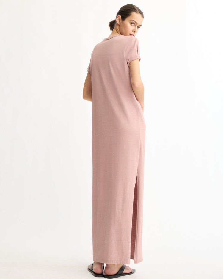 A person faces backward, looking over their shoulder, wearing the Arlo Mott Hubert Short Sleeve Maxi Dress in Dusty Rose with a side slit and black sandals, set against a plain white background.