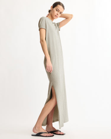 A woman stands sideways against a white background, wearing the Arlo Mott Hubert Short Sleeve Maxi Dress in Grey, ankle-length with a side slit, and black sandals. Made in New York.