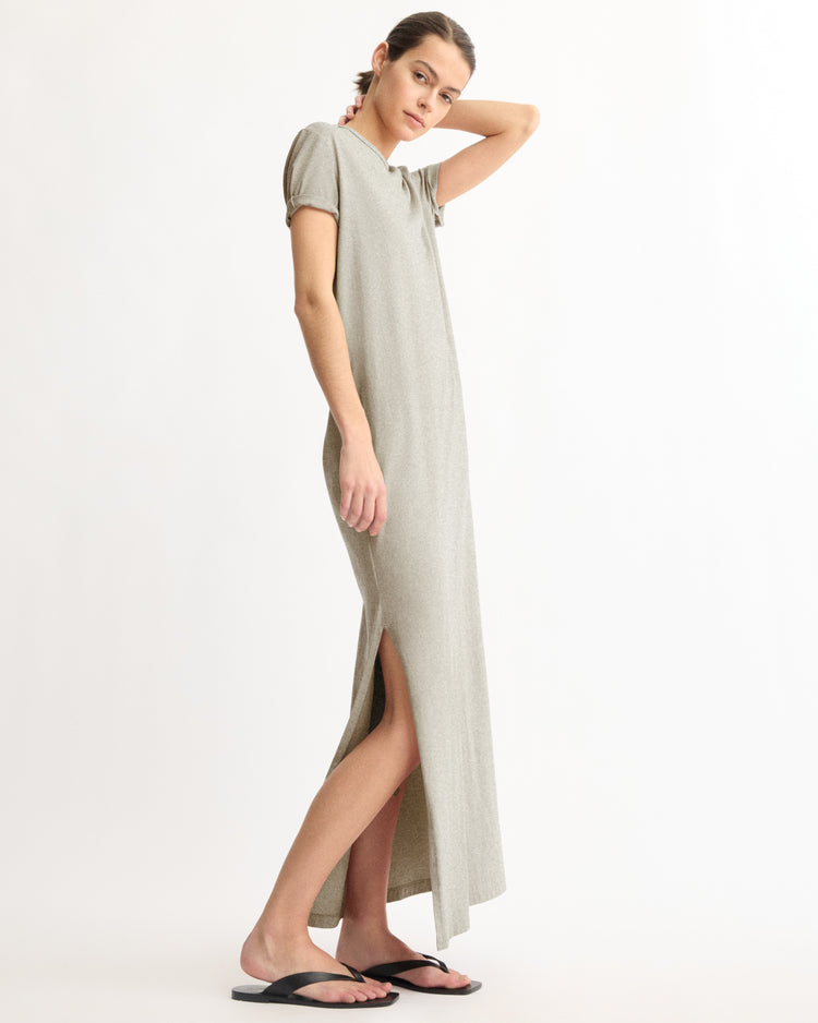 A woman stands sideways against a white background, wearing the Arlo Mott Hubert Short Sleeve Maxi Dress in Grey, ankle-length with a side slit, and black sandals. Made in New York.