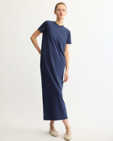 A person stands against a plain background wearing the Arlo Mott Hubert Short Sleeve Maxi Dress in Navy and light-colored flat shoes, with one hand behind their back and a relaxed expression.