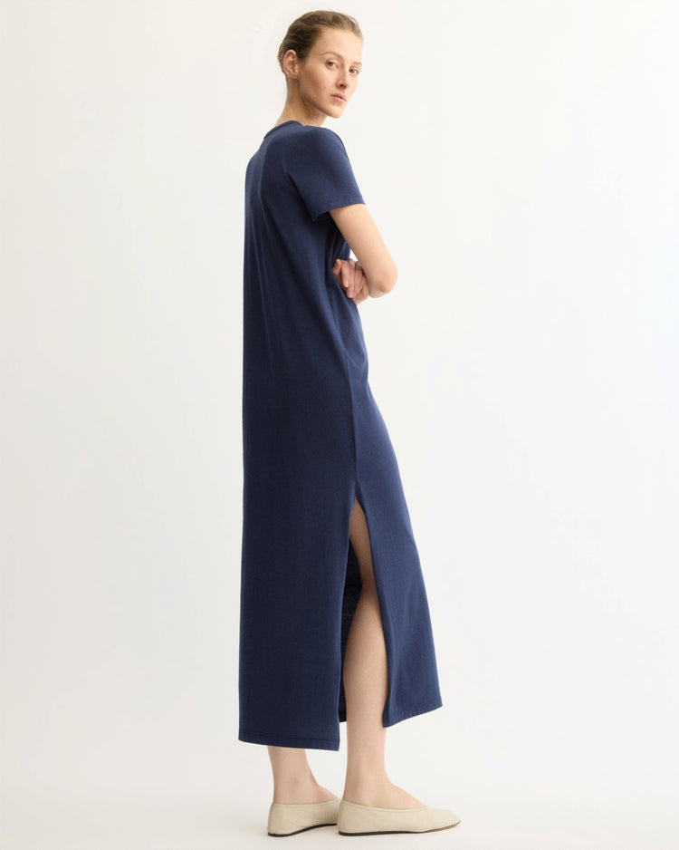 A person stands sideways wearing the Arlo Mott Hubert Short Sleeve Maxi Dress in Navy, made from soft Supima cotton with a high side slit, paired with light slip-on shoes. The background is plain and light-colored.