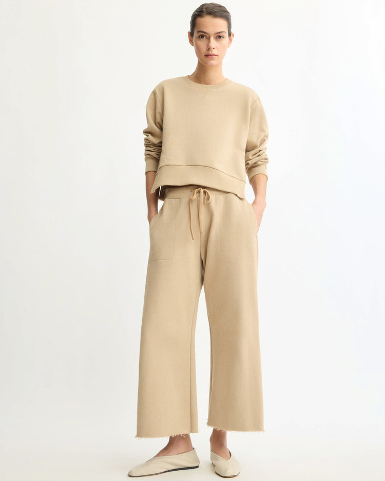 A person stands against a white background, wearing Arlo Mott's Kane Cropped Wide Sweatpants in Khaki with a loose beige Italian terry sweatshirt and beige slip-ons. Made in New York.