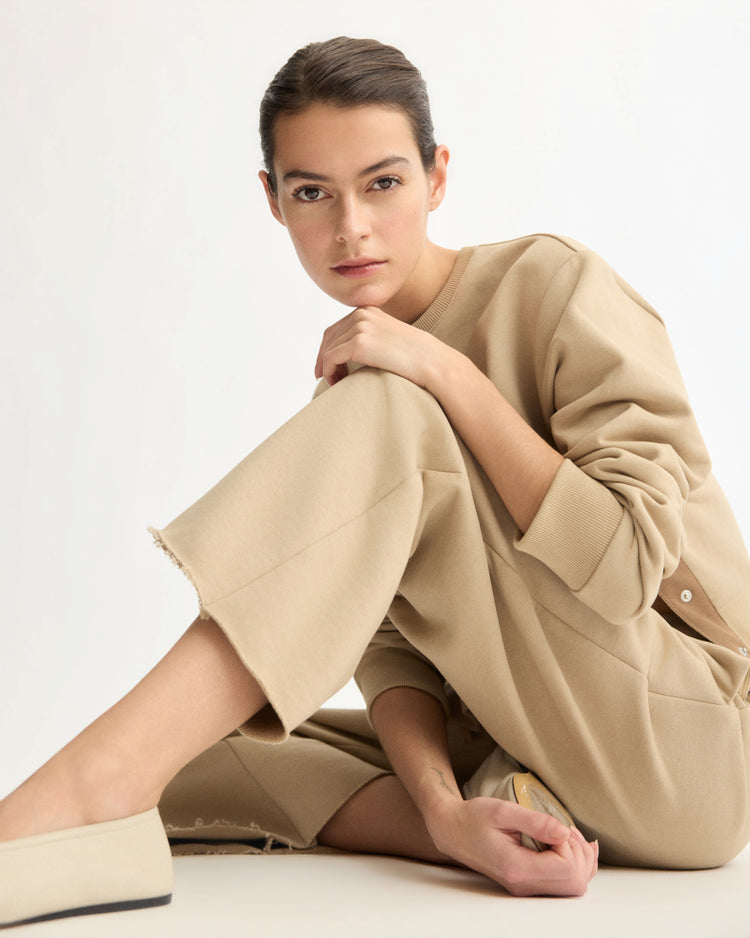 A woman with dark hair pulled back sits on the floor in a beige outfit, featuring Arlo Mott’s Kane Cropped Wide Sweatpants in Khaki, made of Italian terry cloth. She gazes thoughtfully at the camera against a plain, light background.