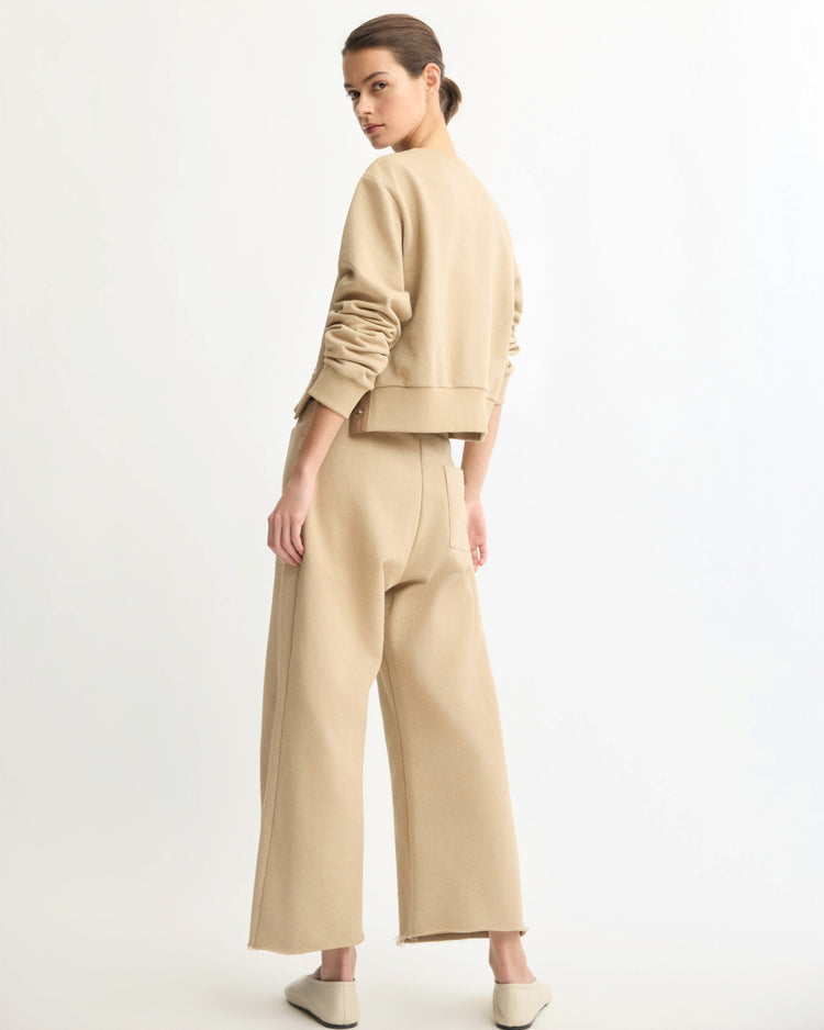 A woman poses against a white background, wearing Arlo Mott's Kane Cropped Wide Sweatpants in Khaki with a beige sweater and flat beige shoes. Her hair is pulled back as she stands facing slightly away.