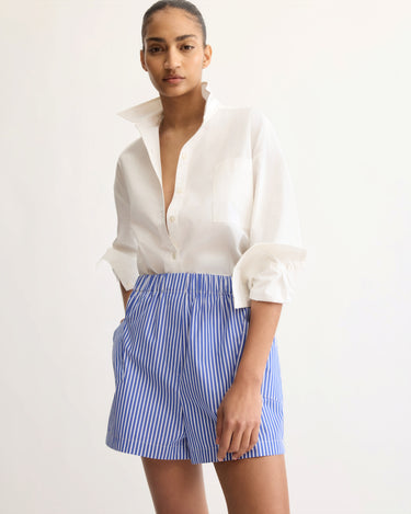 A woman stands against a plain background, confidently looking at the camera in a white button-up shirt with rolled sleeves and Arlo Mott’s Laight Shorts in Blue Stripe, featuring a high rise cut and elastic waistband.