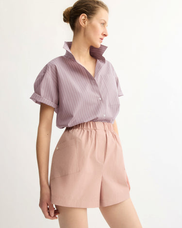 A person stands against a plain light background wearing a short-sleeved, lavender striped button-up with the collar popped and Arlo Mott Laight Shorts in Dusty Rose. Their hair is styled in a bun.