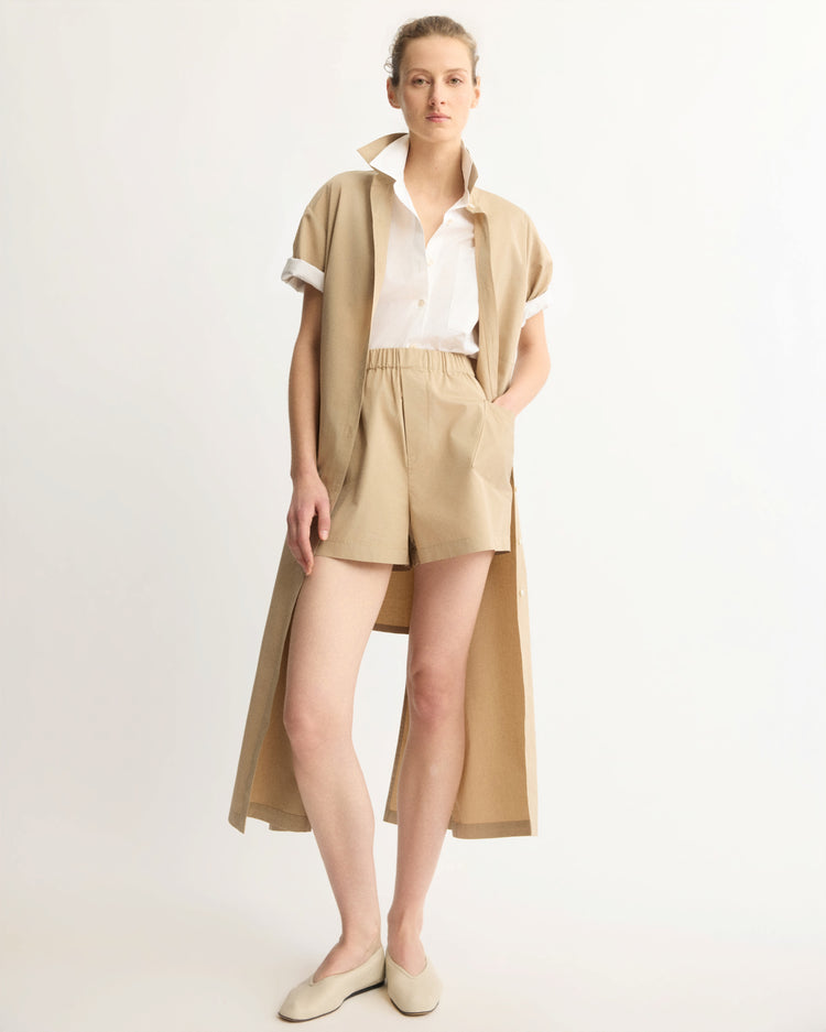 A person stands against a white background wearing Arlo Mott’s Laight Shorts in Khaki with a long beige short-sleeve vest, a white shirt, and beige flats, one hand in their pocket and a relaxed expression.