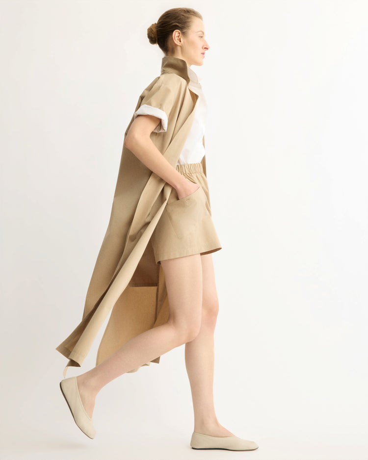 A woman in a beige trench coat, white shirt, and Arlo Mott Laight Shorts in Khaki walks in profile against a plain light background, wearing beige flats and carrying a beige tote bag.