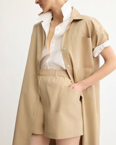 A person wears Arlo Mott’s Laight Shorts in Khaki, styled with a loose beige shirt and a white button-up layered underneath. They stand with one hand in their pocket against a plain background.