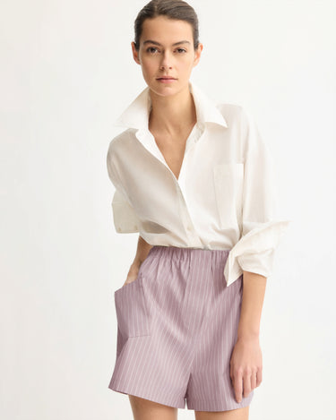 A woman with dark hair tied back wears a loose white button-up shirt and Arlo Mott’s Laight Shorts in Rose Stripe, featuring light purple pinstripes and an elastic waistband, as she poses against a plain light background.