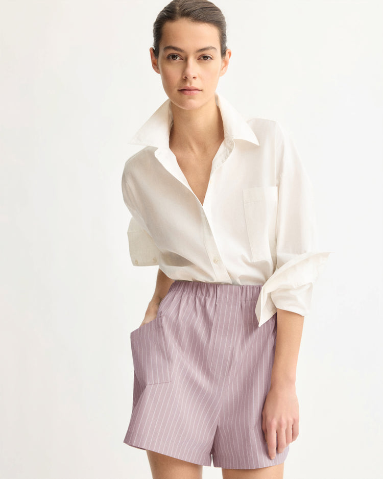 A woman with dark hair tied back wears a loose white button-up shirt and Arlo Mott’s Laight Shorts in Rose Stripe, featuring light purple pinstripes and an elastic waistband, as she poses against a plain light background.