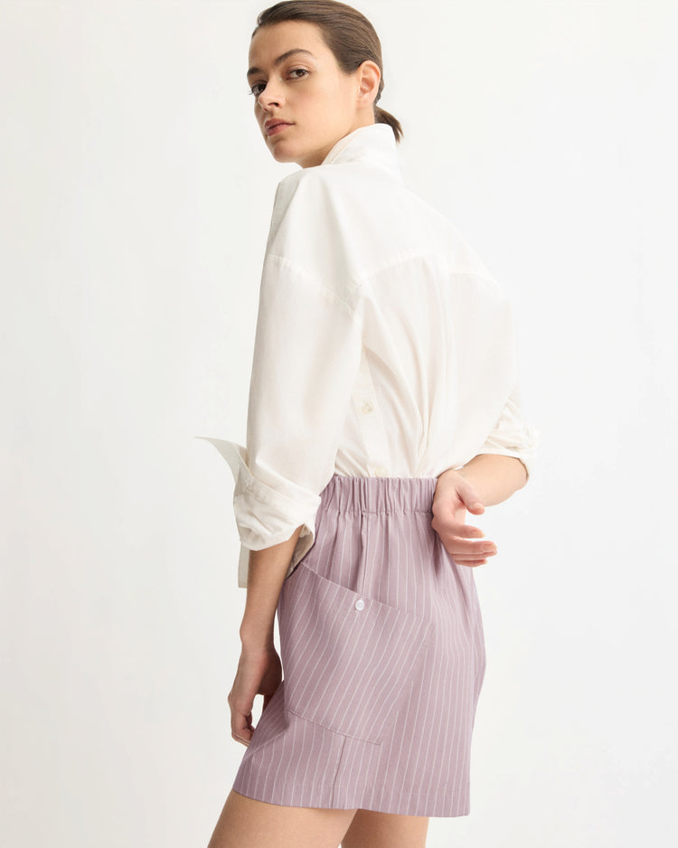 A person stands turned slightly away, looking over their shoulder. They wear a white button-up shirt with rolled sleeves and Arlo Mott's Laight Shorts in Rose Stripe, which have a high rise, elastic waistband, and back pocket against a plain background.