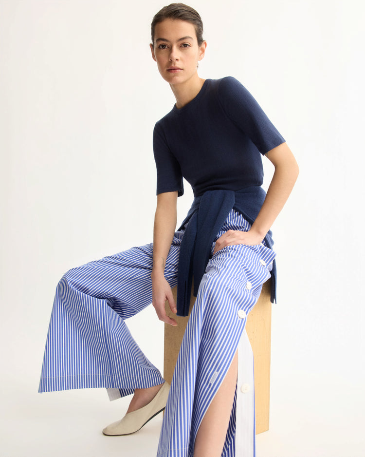 A woman sits on a wooden block wearing a navy short-sleeve top, beige flats, and Arlo Mott’s Baxter Side Button Pants in Blue Stripe with an elastic waist. A navy sweater is tied around her waist as she calmly looks at the camera.