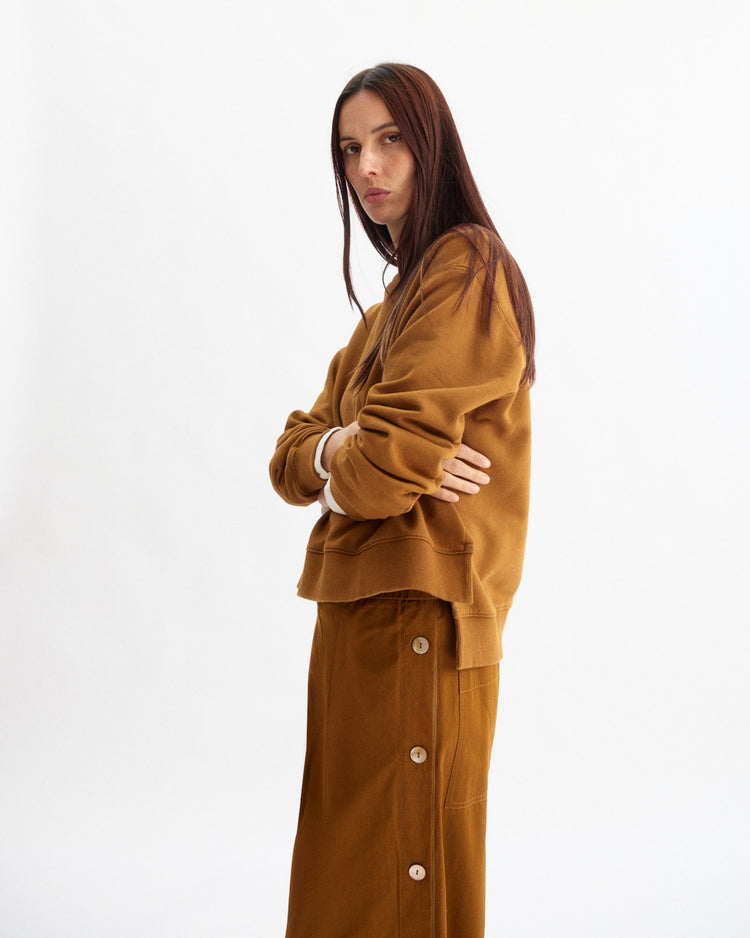 A person with long brown hair stands with arms crossed, wearing a loose brown sweater and Arlo Mott's Baxter Side Button Pants in Cinnamon, made from Portuguese cotton with an elastic pull-on waist, against a plain white background.