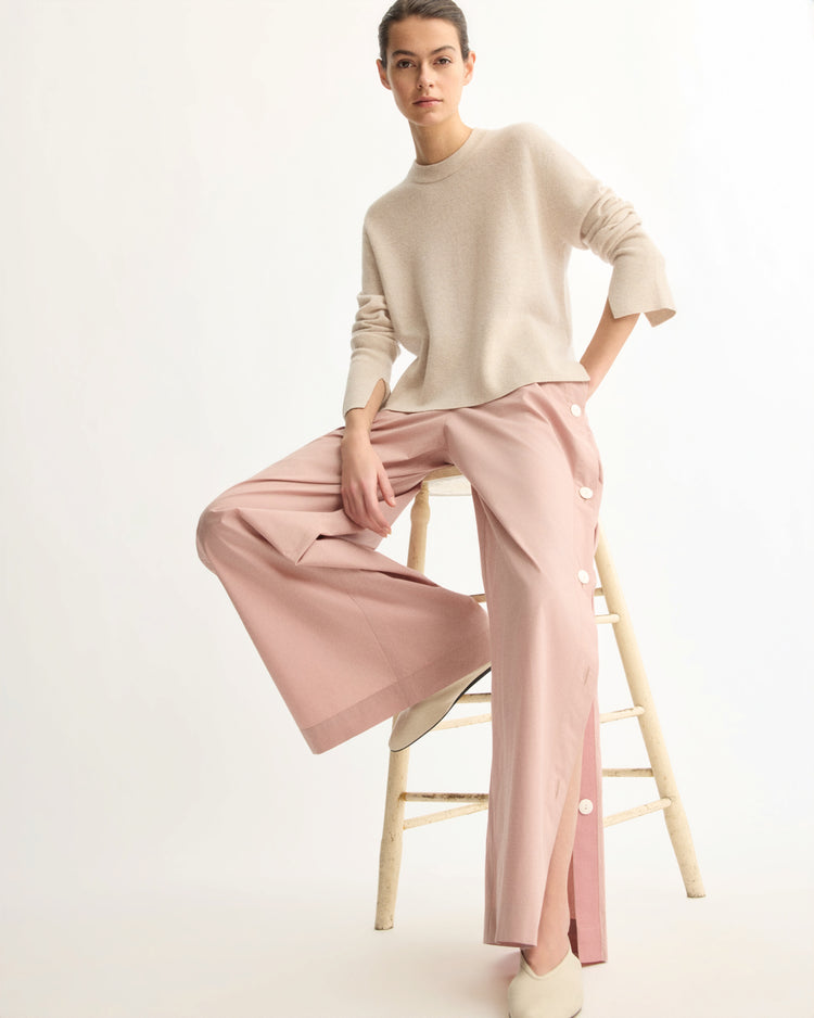 A person with short hair and a relaxed pose sits on a wooden stool against a plain background, wearing a loose beige sweater and Arlo Mott’s Baxter Side Button Pants in Dusty Rose, paired with white slip-on shoes.