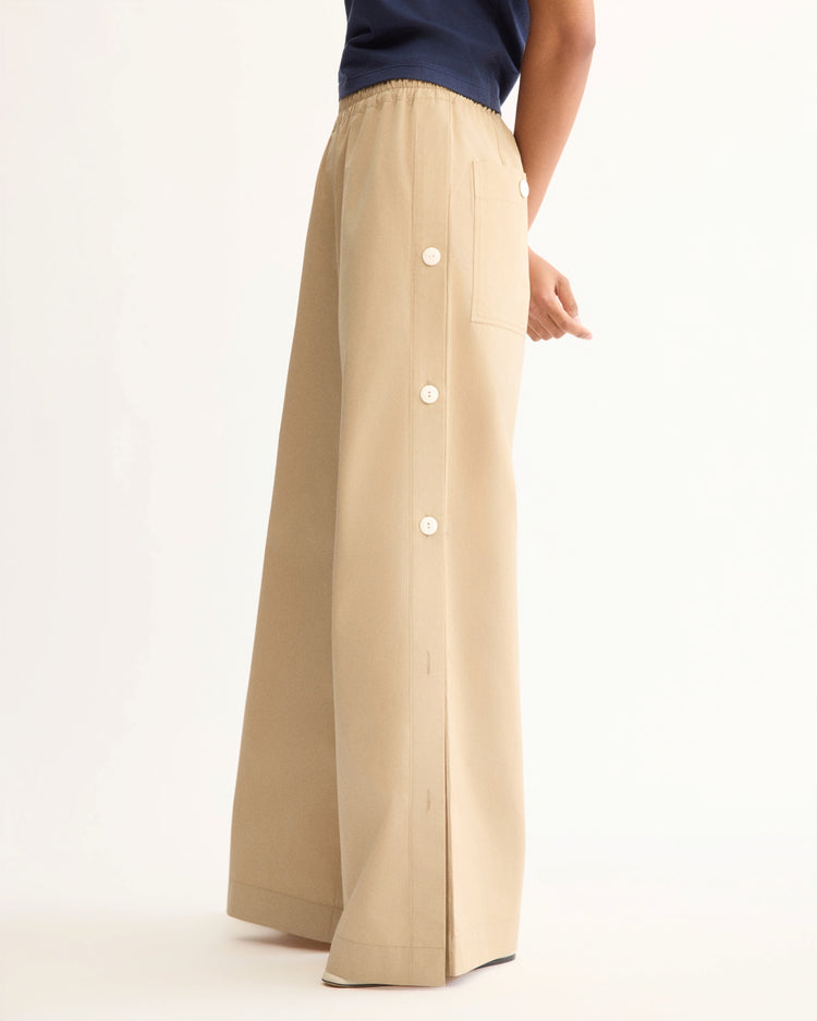 A person wears Arlo Mott's Baxter Side Button Pants in Khaki, featuring an elastic waist, white side buttons, and a large side pocket, paired with a navy top against a plain white background.