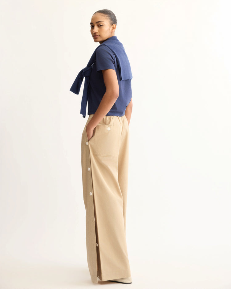 A person stands sideways in a navy blue short-sleeve shirt with another blue top draped over their shoulders, paired with Arlo Mott's Baxter Side Button Pants in Khaki, featuring side buttons, pockets, and an elastic pull-on waist. The background is light and plain.