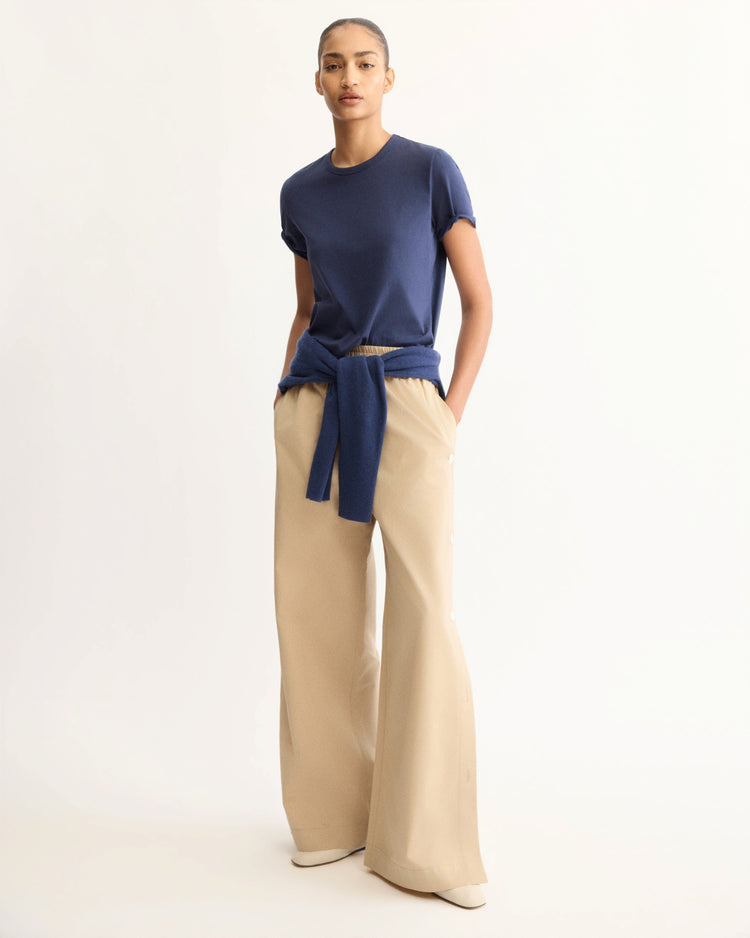 A person stands against a plain background wearing a navy blue t-shirt, Arlo Mott’s Baxter Side Button Pants in Khaki with hands in their pockets, cream shoes, and a blue sweater tied at the waist. Their hair is pulled back.