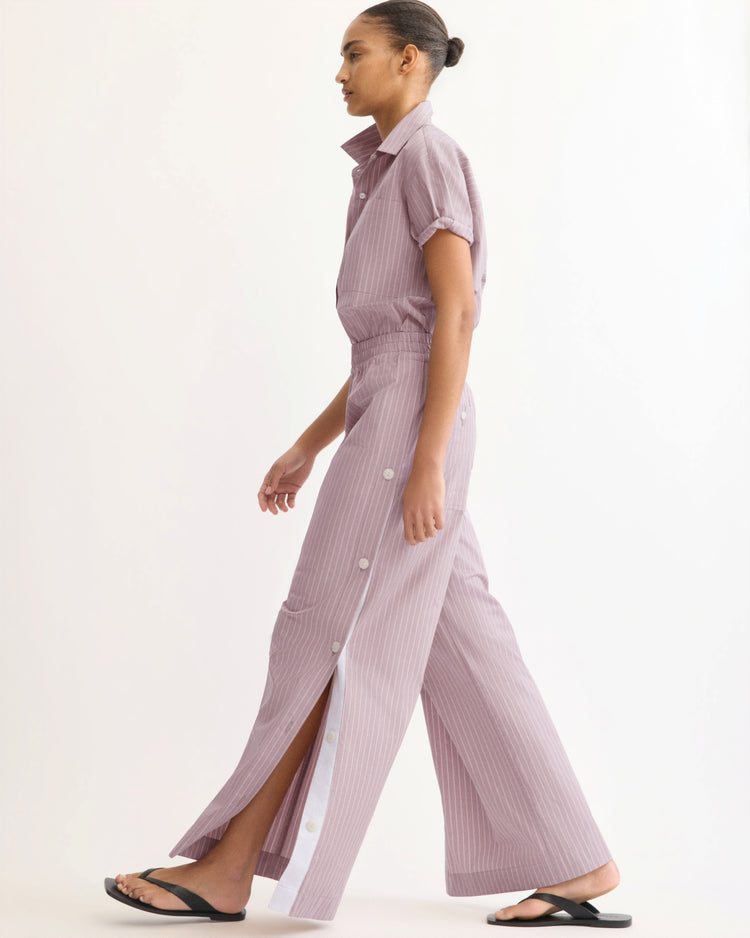 Wearing Arlo Mott’s Baxter Side Button Pants in Rose Stripe with a light purple pinstriped short-sleeve shirt and white flip-flops, a person walks against a plain background.