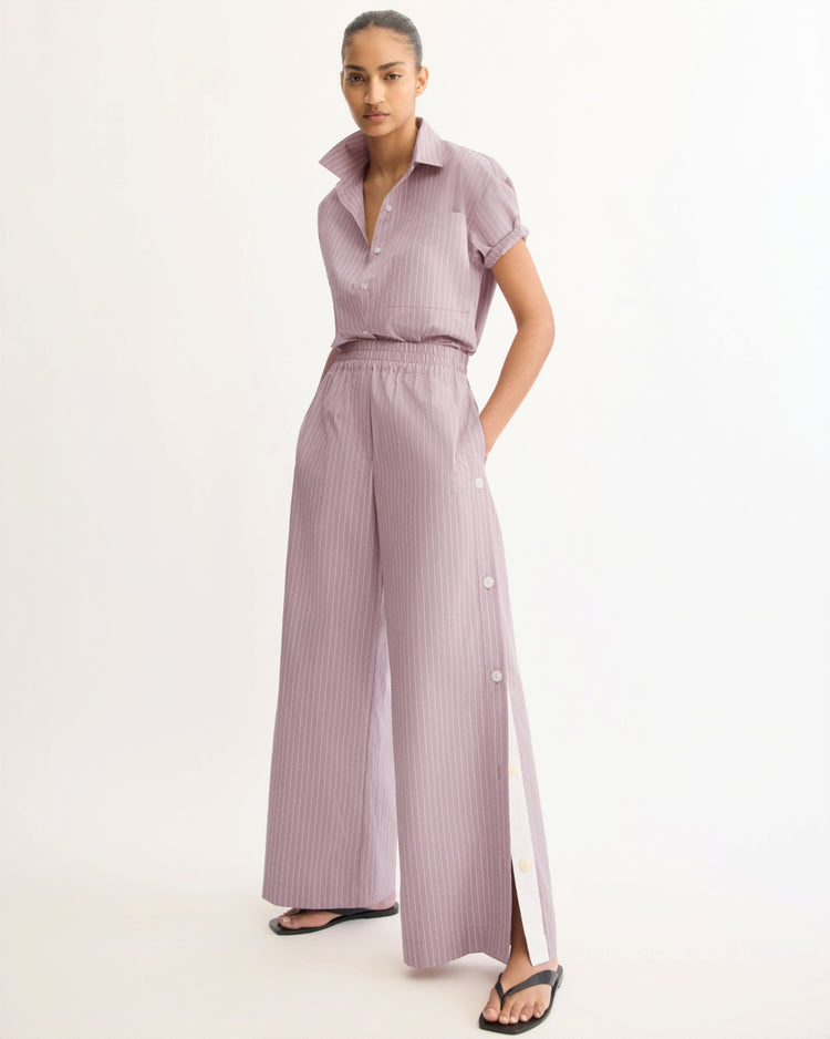 A person poses against a plain background wearing Arlo Mott's Baxter Side Button Pants in Rose Stripe with an elastic waist and side buttons, paired with a matching lavender pinstripe short-sleeve shirt and black sandals.