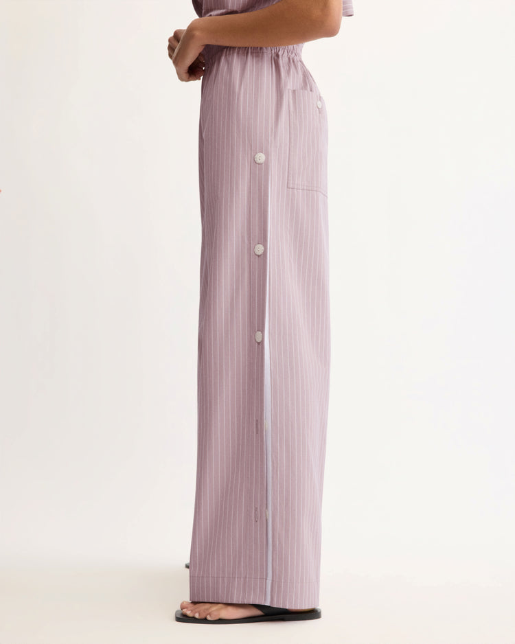 A person stands sideways against a plain white background wearing Arlo Mott’s Baxter Side Button Pants in Rose Stripe—loose-fitting Portuguese cotton pants with lilac pinstripes, an elastic waist, side white buttons—paired with black sandals.