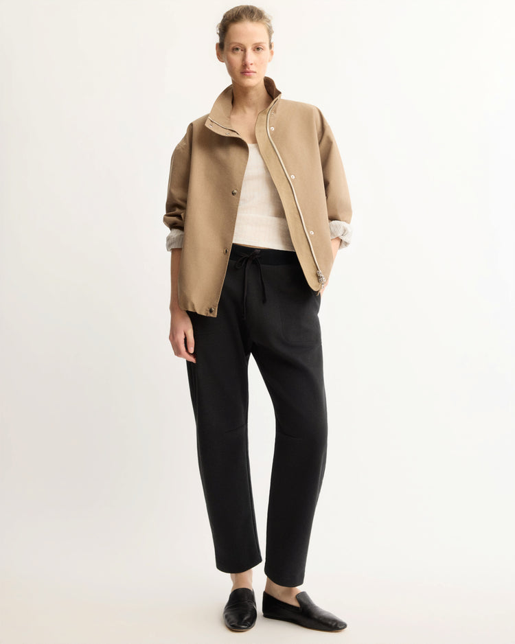A person stands against a plain background wearing Arlo Mott’s Baltic Barrel Leg Sweatpants in Black, a beige jacket over a white top, and black slip-on shoes. Their hair is pulled back and their hands rest at their sides.