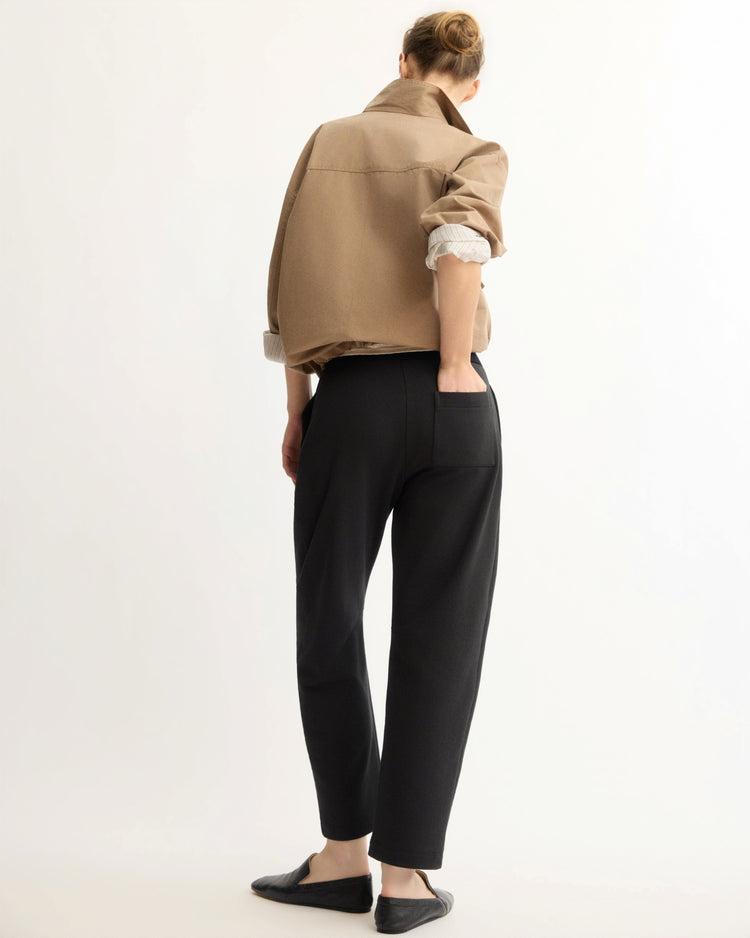 A person faces away from the camera in a tan jacket with rolled sleeves, black flats, and Arlo Mott’s Baltic Barrel Leg Sweatpants in Black, set against a plain light background.