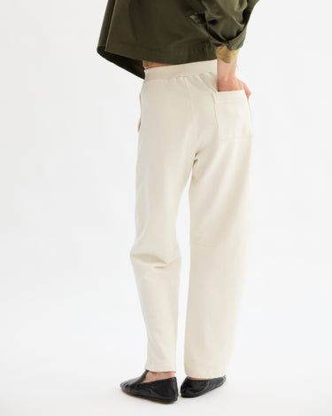 A person stands with their back turned, wearing Arlo Mott's Baltic Barrel Leg Sweatpants in Chalk, paired with a dark green top and black flats, one hand in the back pocket, against a plain white background.