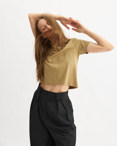A woman with long hair wears the Arlo Mott Grand Wide Leg Pant in Black and a tan crop top, posing with her arms raised and eyes closed against a plain white background.
