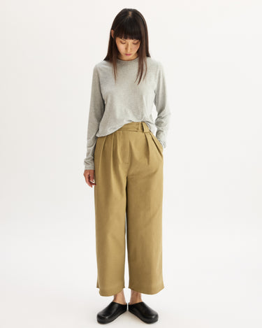 A woman stands against a white background, wearing a light gray long-sleeve top, Arlo Mott’s Grand Wide Leg Pant in Khaki, and black shoes. She looks down with her hands in her pockets.