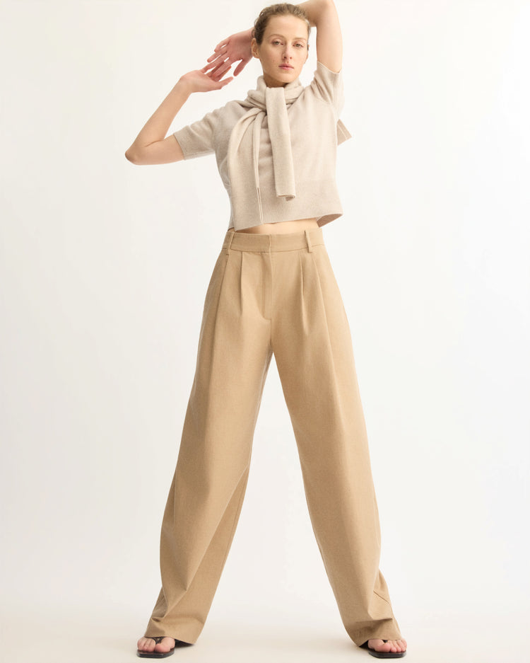 A woman poses with one arm raised above her head, wearing Arlo Mott's Horatio Pleated Barrel Trouser in Khaki with a wide-leg fit, paired with a cropped beige sweater and a matching sweater draped over her shoulders.