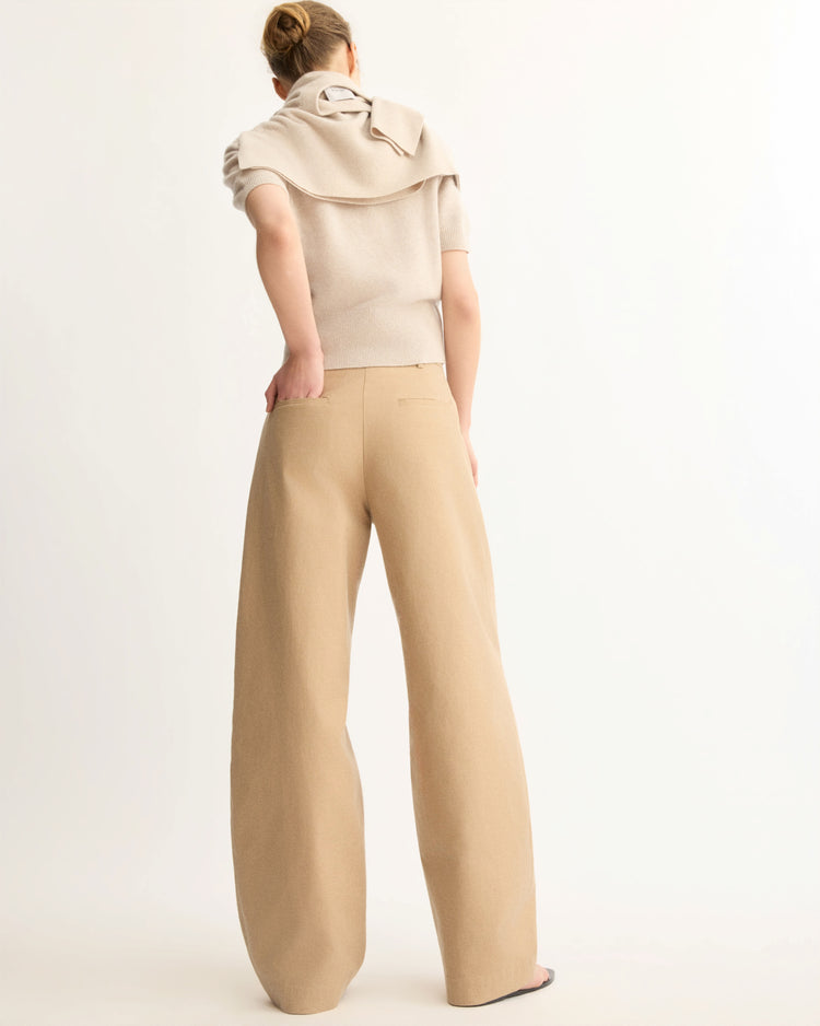 A person faces away, wearing a beige short-sleeve top with a matching sweater over their shoulders and Arlo Mott's Horatio Pleated Barrel Trouser in Khaki. Their hair is tied back and the background is plain white.