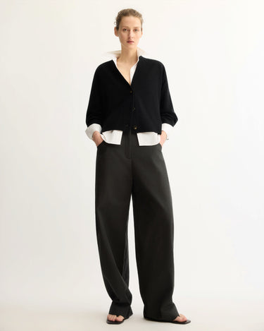 A person stands against a plain light background, wearing a black buttoned cardigan over a white collared shirt, Arlo Mott’s Leroy Flat Front Barrel Pant in Black, and black sandals, with hands in pockets in a relaxed pose.