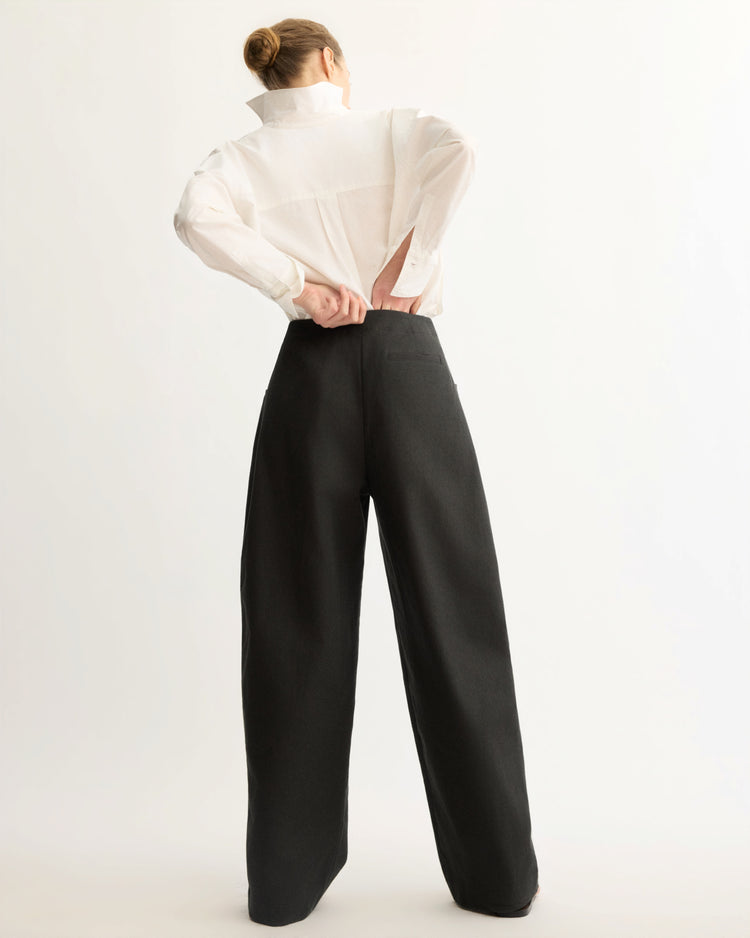 A person stands facing away against a plain light background, wearing a white shirt and Arlo Mott's Leroy Flat Front Barrel Pant in Black, featuring a high-waisted barrel silhouette in Japanese twill.