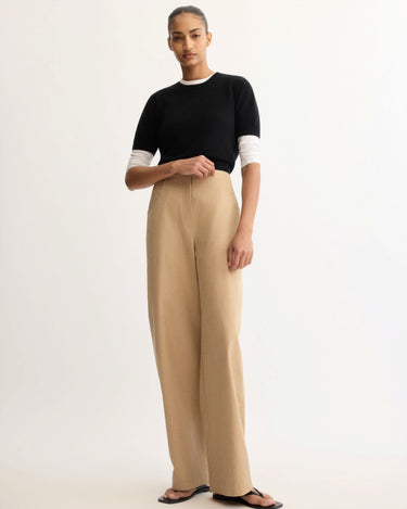 A person stands against a light background wearing a black short-sleeve top over a white long-sleeve shirt, Arlo Mott Leroy Flat Front Barrel Pant in Khaki, and black sandals. Their hair is pulled back with a neutral expression.