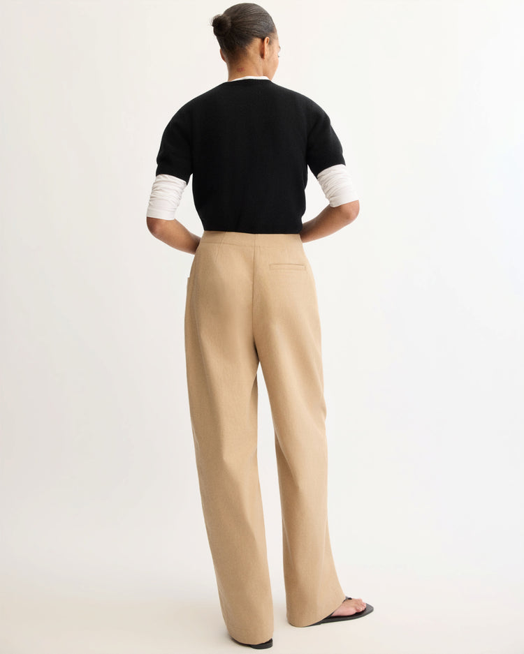 A person with dark hair in a bun faces away, wearing a black short-sleeve sweater over a white long-sleeve top and Arlo Mott Leroy Flat Front Barrel Pant in Khaki, paired with black sandals against a plain light background.
