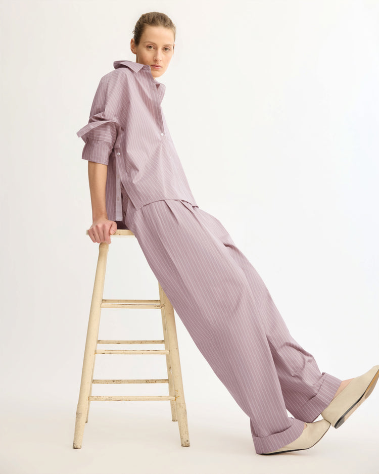 A person in Arlo Mott's Spencer Pleated Pants in Rose Stripe leans back on a wooden ladder against a plain white background, paired with a loose-fitting pinstripe shirt for a coordinated look.