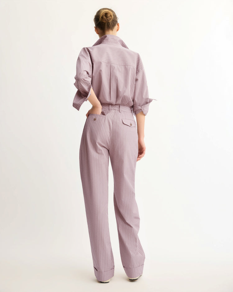 A person with a bun faces away, wearing Arlo Mott's Spencer Pleated Pants in Rose Stripe—light purple pinstripes, high-waisted with rolled cuffs—paired with a matching long-sleeve shirt, against a plain white background.